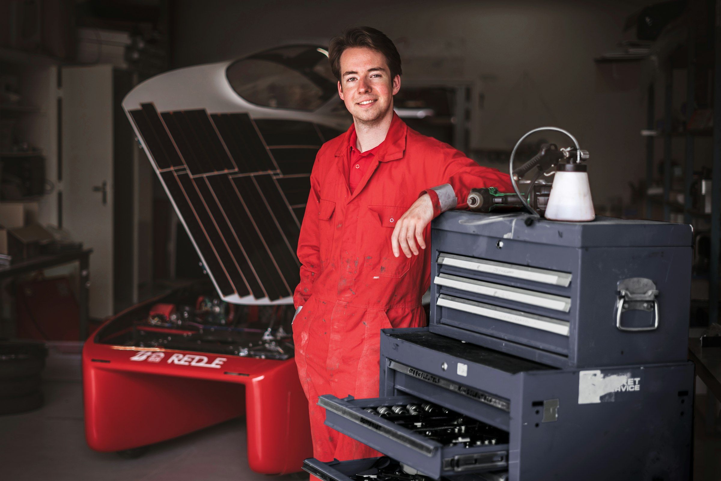 Mecánico sonriente con mono rojo apoyado en su caja de herramientas en el taller